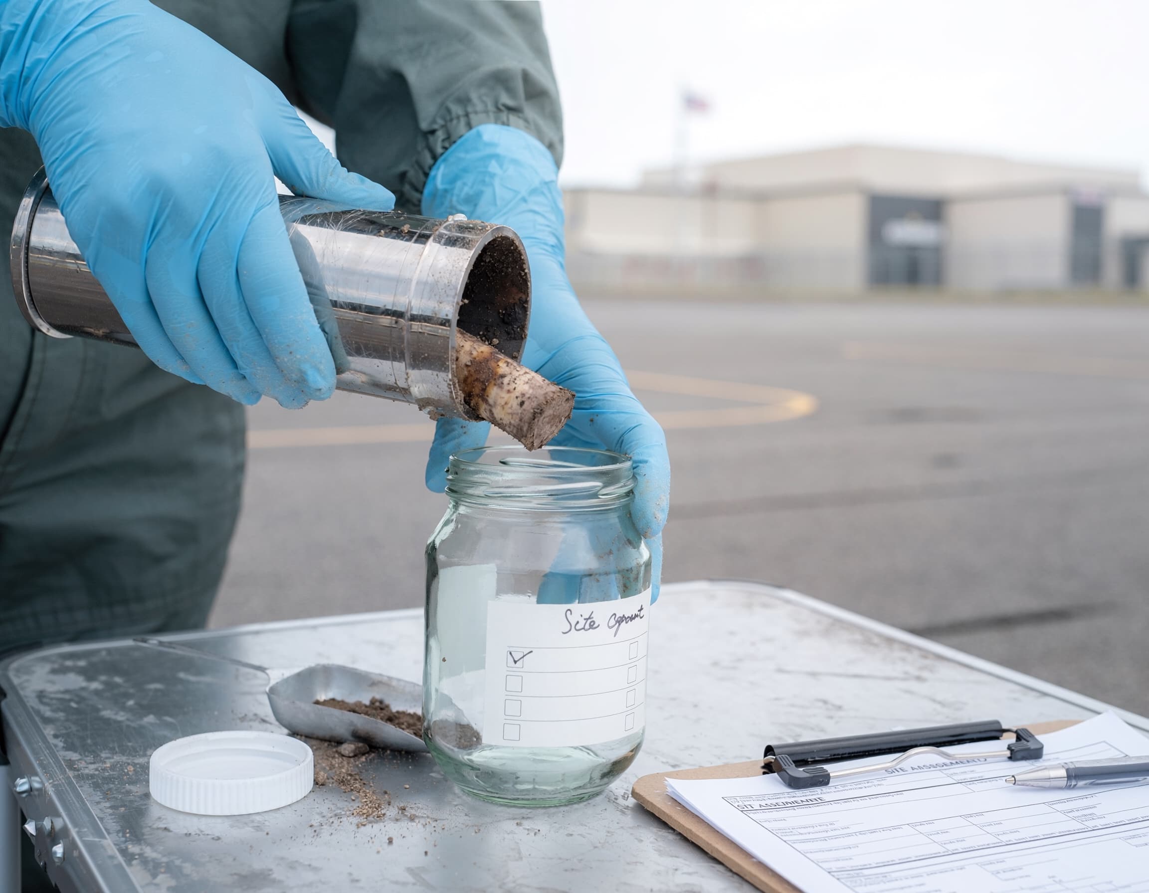 Gloved hands extracting a soil core sample into a labeled glass jar at a federal facility.